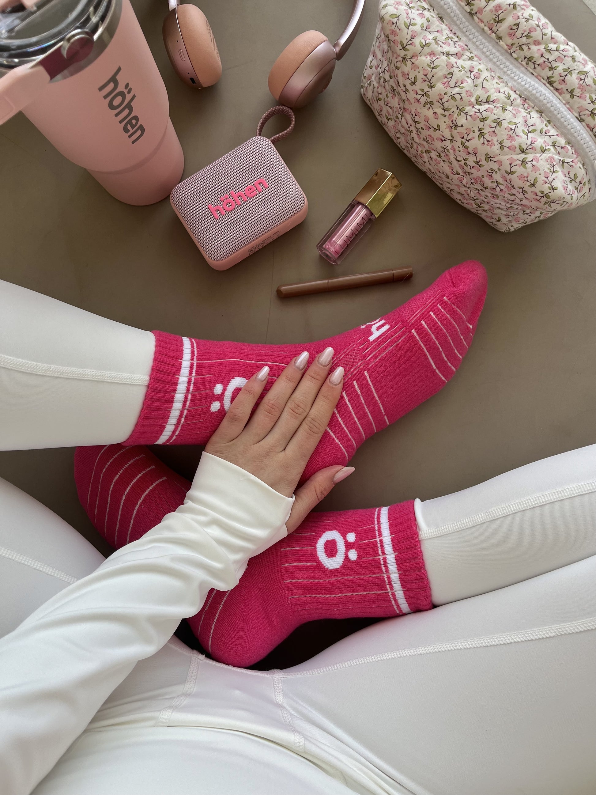Top-down lifestyle view of a woman wearing magenta höhen core athletic socks, surrounded by a matching pink höhen essential tumbler, höhen move wireless portable speaker, and höhen aura mini headphones.