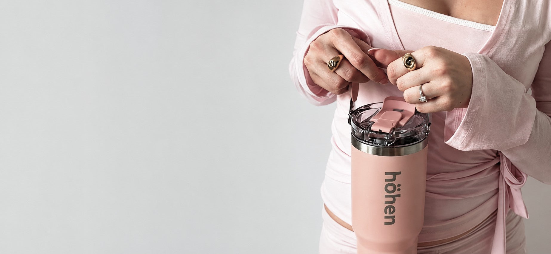A close-up shot of a person wearing a light pink long-sleeve top and matching bottoms, holding a pink "höhen" insulated stainless steel tumbler. The person's hands are decorated with large gold rings and a silver ring. The tumbler features a transparent plastic lid with a pink flip-top straw mechanism and a built-in carrying handle. The brand name "höhen" is printed vertically in grey letters on the front of the matte pink tumbler body.
